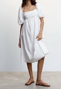 Weißes Kleid mit Puffärmeln, quadratischem Ausschnitt, gerafftem Oberteil und fließendem Rock. Kombiniert mit flachen braunen Sandalen. Glatte Stoffstruktur.