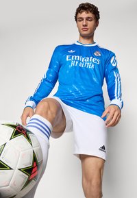 Male soccer player in blue goalkeeper jersey and white shorts controlling soccer ball with his knee against white background.