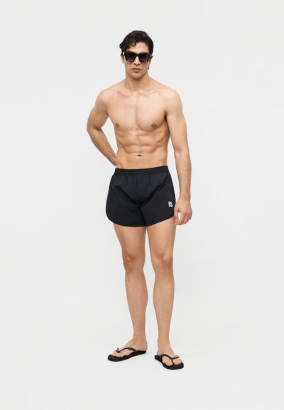 Άνδρας χωρίς πουκάμισο που φοράει μαύρα μαγιό, μαύρες σαγιονάρες και μαύρα γυαλιά ηλίου, στέκεται μπροστά σε απλό φωτεινό φόντο.