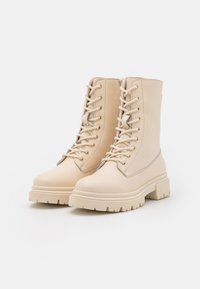 Beżowe botki do kostek z gładkiej skóry, z frontem sznurowanym, gumową podeszwą i masywnym bieżnikiem, z złotymi oczkami i detalami w kolorze złotym.