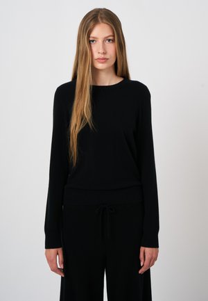 Jonge vrouw met lang, stijl haar, die een effen zwarte top met lange mouwen en een zwarte broek draagt tegen een lichtgrijze achtergrond.