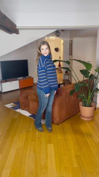 Suéter às riscas azuis com mangas largas, detalhes em azul claro e corte descontraído; combinado com jeans azuis ajustados. O fundo apresenta um sofá castanho.