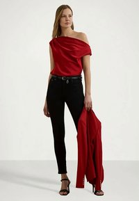 Žena v červenom off-shoulder blúzke, čiernych nohaviciach a čiernych lodičkách, drží červený sako v pravej ruke a stojí na neutrálnom pozadí.