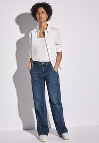 Blå denimbyxor med vida ben och sidostuddetaljer, tillsammans med en vit tanktop och en vit skjorta, samt vita sneakers.