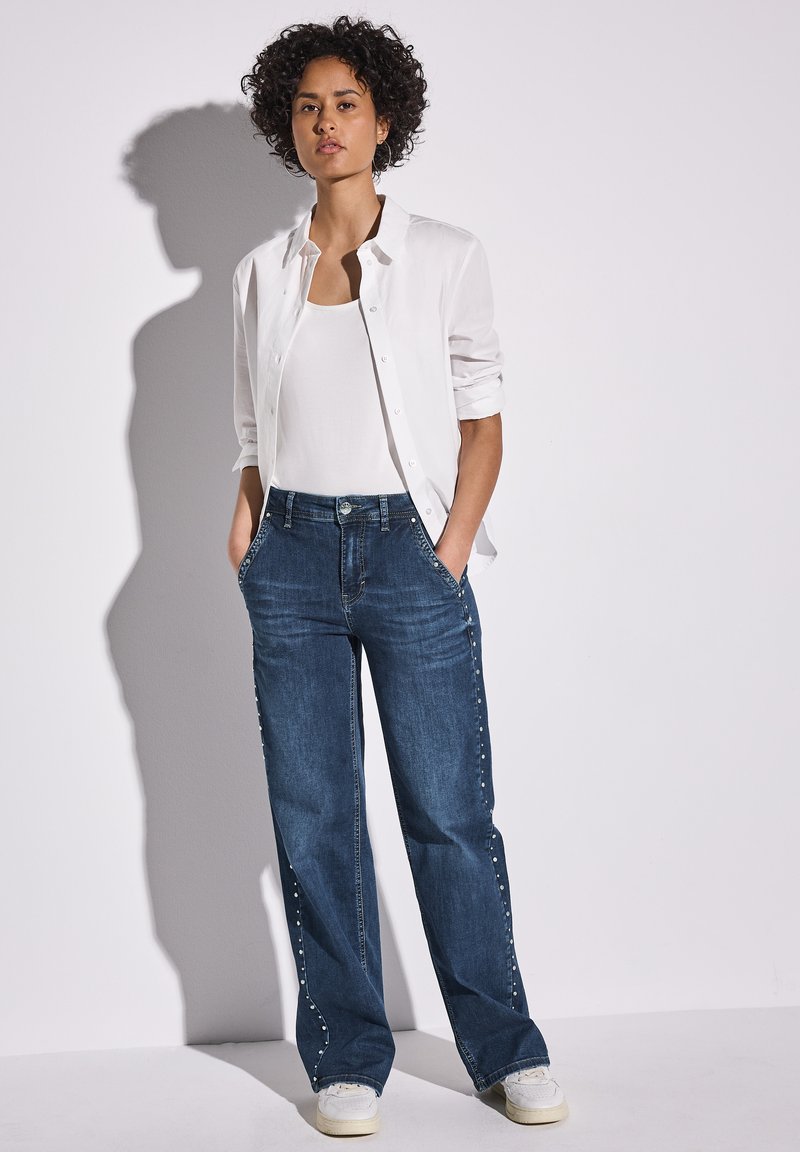 Blå denimbyxor med vida ben och sidostuddetaljer, tillsammans med en vit tanktop och en vit skjorta, samt vita sneakers.