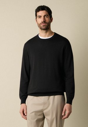 Man staat en draagt een zwart trui met lange mouwen over een wit shirt en beige broek tegen een effen beige achtergrond.