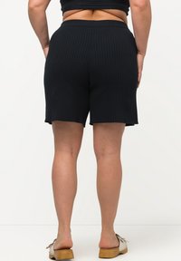 Mustat ribattu shortsit, joissa on istuva vyötärö, polven ylle ulottuva pituus, teksturoitu kangas ja aaltoileva helma. Yhdistetty puisiin korkokenkiin.