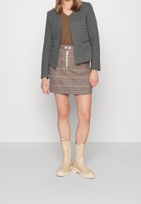 Blazer gris ajusté, chemise marron à col en V, jupe marron à motif pied-de-poule avec cordon de serrage, et bottes beige montant jusqu'aux genoux avec une semelle épaisse.
