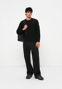 Pulover texturat, negru, cu guler rotund, asortat cu pantaloni largi negri. Persoana poartă un rucsac negru. Încălțămintea este sportivă și de culoare închisă.