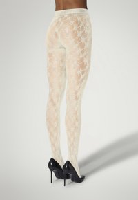 Weiße Spitze-Strumpfhosen mit einem floralen Muster, semi-transparent, kombiniert mit schwarzen High Heels. Elastischer Bund, strukturierte Oberfläche.
