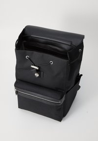 Sac à dos en tissu noir avec une fermeture à cordon, poche avant zippée et accents noirs brillants. Il présente un design structuré et des poignées robustes.
