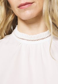Gros plan sur le bas du visage et le cou d'une femme portant un chemisier blanc avec un col haut plissé orné de dentelle.