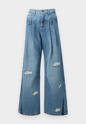 Kvinde iført en blå stribet blomstrede kortærmet peplumbluse, vidbenede revnede jeans og brune sandaler med åben tå, stående mod hvid baggrund.