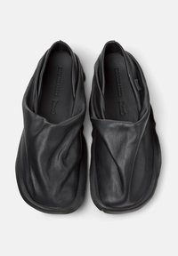 Scarpe slip-on in pelle nera con design drappeggiato, punta rotonda e texture liscia. Presentano un marchio minimalista sulla soletta.