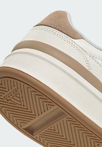 Zapatilla blanca con acentos de ante beige. Presenta una parte superior de cuero texturizado y una suela de goma con un patrón de espiga. Diseño deportivo.