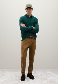 Junger Mann mit verschränkten Armen, bekleidet mit einem grünen Langarm-Polohemd, braunen Hosen, einem geflochtenen Gürtel, schwarzen Schuhen und einer gemusterten Mütze.