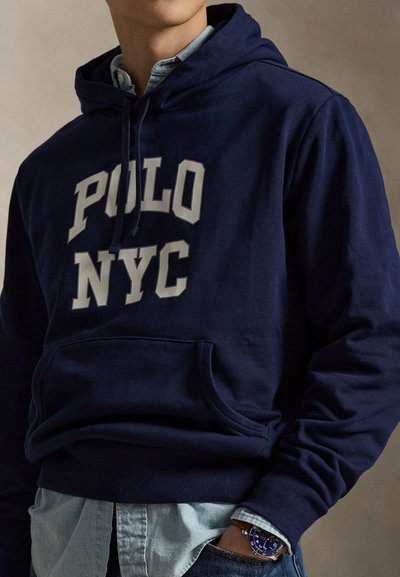 Μπλε σκούρα φούτερ με κουκούλα και το γράφημα «POLO NYC» σε γκρι στο μπροστινό μέρος. Διαθέτει τσέπη καγκουρό και φοριέται πάνω από ένα ανοιχτό μπλε πουκάμισο με γιακά.