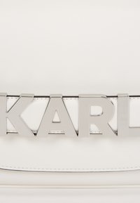 Biała skórzana torebka z błyszczącym srebrnym logo "KARL", o teksturowanym wykończeniu i prostych liniach, uzupełniona czarnymi przeszyciami.