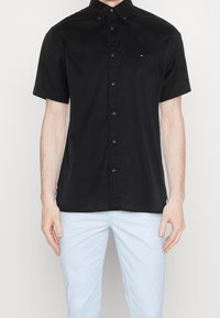 Man in een zwart overhemd met korte mouwen en lichte blauwe broek, staand tegen een effen achtergrond.