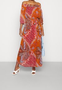 Kvinna som bär en orange och röd mönstrad off-shoulder maxiklänning med knytning i midjan, vita klacksandaler och bär en ljusblå handväska.