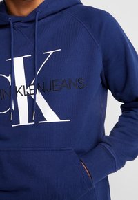 Felpa blu navy in cotone, con un grande logo bianco 'CK' e il testo nero 'KLEINJEANS' sul davanti. Include cordini e tasca.
