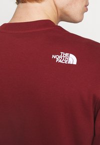 Persona che indossa un maglione girocollo bordeaux con il logo bianco "The North Face" sulla parte superiore della schiena, mostrando parzialmente il profilo laterale del viso.