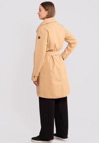 Vedenkestävä beige takki, jossa on vyötärölle kiinnitettävä vyö, nappikäänteet ja suuri kaulus. Vasemman hihan ylle on ommeltu logopatch.