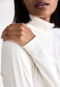 Ribbed, wysoka bluzka w kolorze écru z długimi rękawami. Zbliżenie na dłoń spoczywającą na ramieniu, ukazujące strukturę skóry i detale materiału.