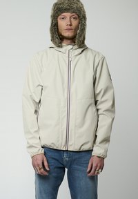 Veste imperméable beige clair avec une fermeture éclair à l'avant, des rayures de couleur contrastée et deux poches latérales. Elle est dotée d'une capuche doublée en fausse fourrure.