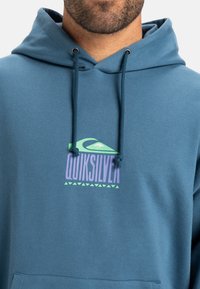 Sweatshirt à capuche bleu avec un logo graphique et des accents verts et violets. Fabriqué en tissu doux, il comprend une poche kangourou et des cordons de serrage.