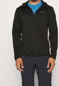 Man som bär en svart Columbia zip-up jacka över en blå skjorta och mörka byxor, står med händerna avslappnat vid sidorna.