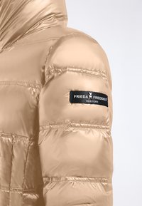 Jasno beżowa kurtka puchowa o błyszczącej, gładkiej fakturze, z poziomymi przeszyciami i czarną metką z logo na ramieniu.