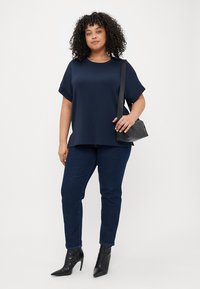 Tamno plavi majica s kratkim rukavima, opuštenog kroja i okruglim izrezom, u kombinaciji s tamnim trapericama i crnim čizmama do članaka. Crna torba preko tijela.