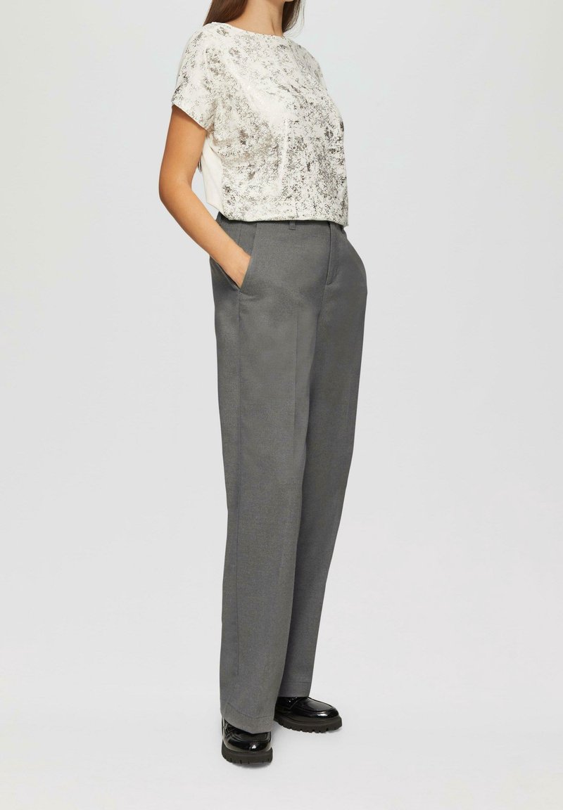 Femme portant un pantalon gris à jambes larges, un haut léger à manches courtes à motif et des chaussures noires brillantes, debout les mains dans les poches.