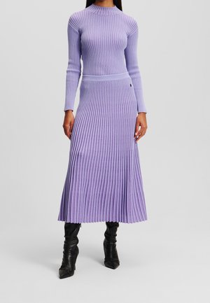 Femme portant une robe en tricot lavande à manches longues et côtes, associée à des bottes noires à bout pointu mi-mollet sur un fond uni.