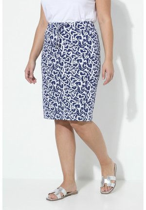 Mujer con falda azul con patrones de corazones blancos, top blanco y sandalias planas plateadas, de pie frente a un fondo blanco liso.