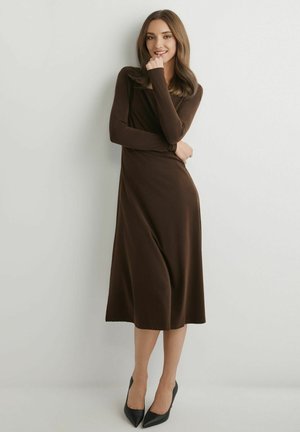 Kobieta w brązowej sukience midi z długim rękawem i czarnych szpilkach stoi przy białej ścianie, uśmiecha się, trzymając dłoń blisko podbródka.