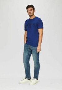 Blue short-sleeve t-shirt with black logo text, paired with light blue jeans and white sneakers. Plain background focuses on the outfit.