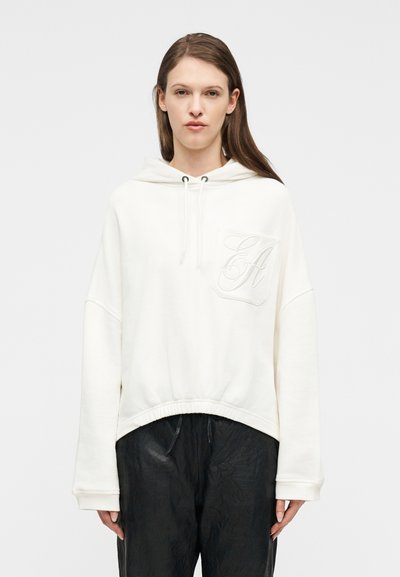 Femme portant un sweat à capuche blanc avec une poche avant ornée d'une broderie cursive "EA" et un pantalon ample noir texturé.