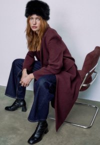 Mørker�ød uldfrakke, blå vide denimbukser, sort faux fur hat og sorte hæle støvler. Siddende pose med en moderne krom stol som baggrund.