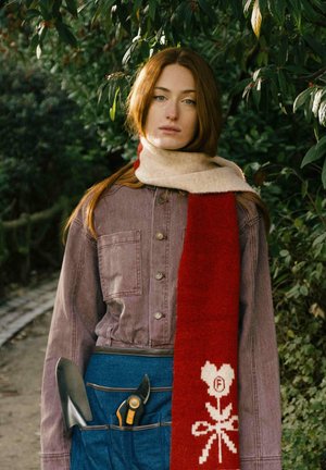 Jeune femme aux longs cheveux roux portant une écharpe rouge et beige, une veste violette et un tablier bleu, tenant des outils de jardinage, debout au milieu d'un feuillage vert.