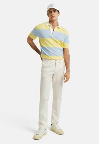 Poloshirt met korte mouwen in geel en blauwe strepen, witte kraag; gecombineerd met witte broek en sneakers met groene accenten.