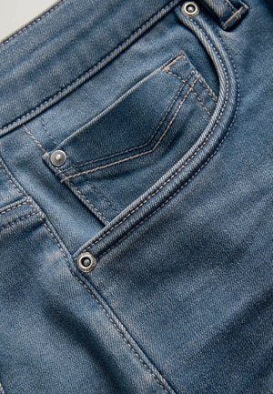 Niebieskie dżinsy wykonane z denimu, z przednią kieszenią zapinaną na srebrny guzik. Tkanina ma wyblakłą fakturę z kontrastującymi przeszyciami.