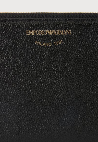 Μαύρη υφασμάτινη δερμάτινη επιφάνεια με χρυστό φερμουάρ και χρυσό λογότυπο Emporio Armani, συμπεριλαμβανομένου του κειμένου "Milano 1981" κάτω από το όνομα της μάρκας.