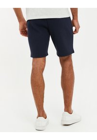 Shorts texturés bleu marine au design ajusté, dotés d'une taille lisse et d'une coupe jusqu'aux genoux, associés à des chaussures blanches à enfiler.