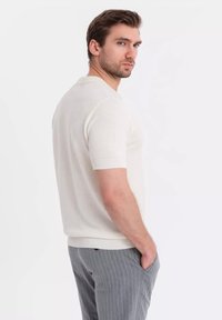 Bílý pletený polokošile s krátkým rukávem a texturovaným vzorem, s rebrovaným límcem a lemem, v kombinaci se světle šedými kalhotami s jemným pruhováním.