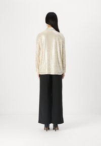 Top beige con paillettes e maniche lunghe, caratterizzato da una vestibilità comoda e colletto a revers. Abbinato a pantaloni neri a gamba larga e tacchi.
