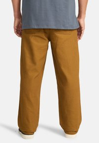 Pantaloni larghi marroni con una finitura testurizzata, caratterizzati da tasche posteriori e dettagli di cucitura visibili, abbinati a scarpe navy.