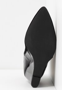 Gros plan sur une semelle de chaussure noire pointue marquée taille 37, pressée contre un mur et un sol blancs, montrant une surface lisse et légèrement texturée.