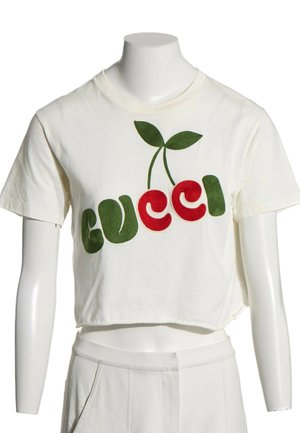 Biała krótka koszulka z zielono-czerwonym wzorem wiśni tworzącym słowo Gucci na białym torsie manekina.